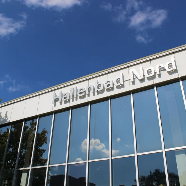 Ein modernes Gebäude mit großen Fenstern reflektiert den blauen Himmel und einige Wolken. Oben am Gebäude prangt der Schriftzug "Hallenbad Nord". Neben dem Gebäude sind einige Bäume zu sehen.