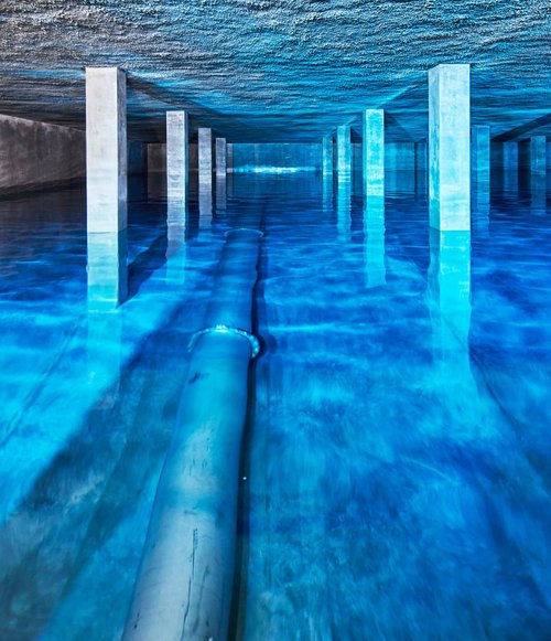 Innenansicht des Trinkwasserbehälters auf dem Sand mit spiegelglatter Wasseroberfläche, Betonstützen und indirektem blauen Licht.
