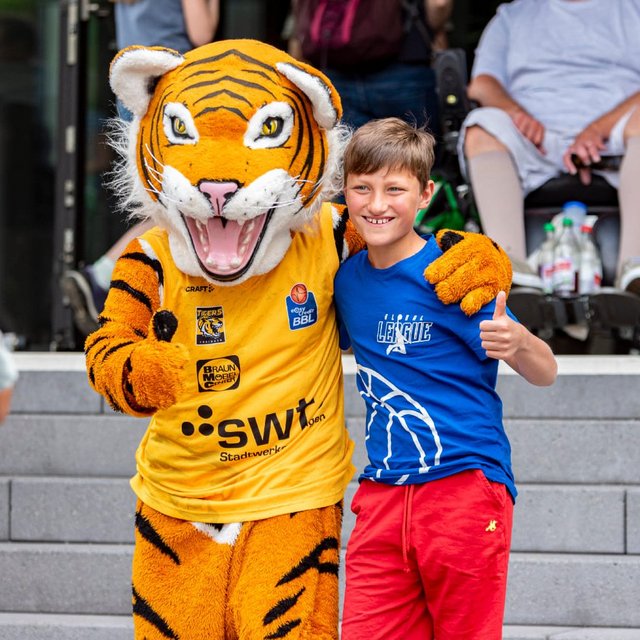 Ein Junge steht neben einem Tiger-Maskottchen, beide lächeln und posieren für ein Foto. Im Hintergrund sind Zuschauer sichtbar