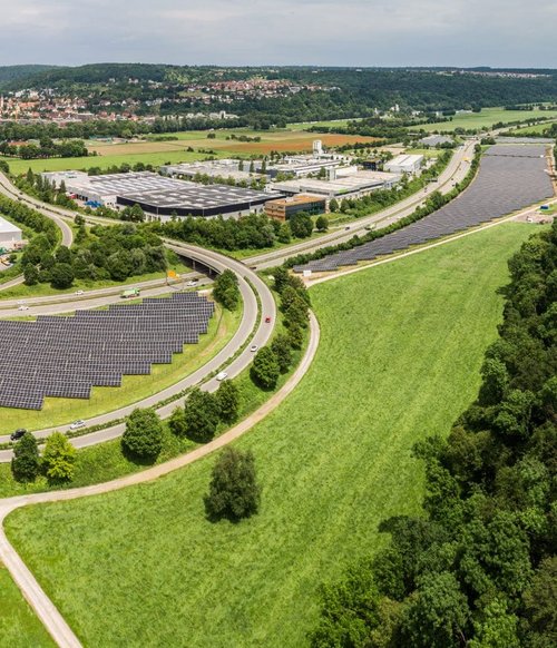 Eine Luftaufnahme zeigt eine weitläufige Landschaft mit einem klaren Himmel. Im Vordergrund sind große Solarpanel-Felder zu sehen, die in einer geschwungenen Form angeordnet sind. Umgeben sind sie von grünen Wiesen und Bäumen. Im Hintergrund sind mehrere moderne Gebäude und Industrieanlagen zu erkennen. Eine Straße schlängelt sich um die Solarpanels und führt zu weiteren Gebäuden. Am Horizont sind Hügel und eine weitere Siedlung sichtbar. Das gesamte Bild vermittelt den Eindruck einer modernen, umweltfreundlichen Infrastruktur.