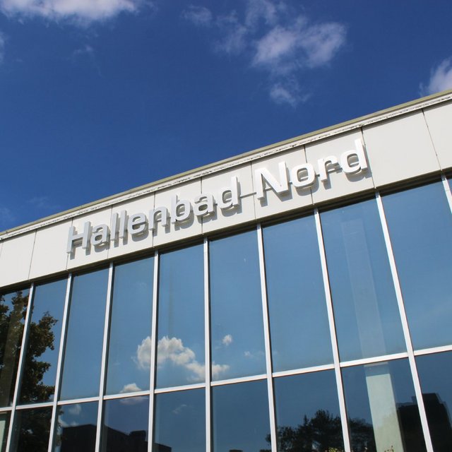 Ein modernes Gebäude mit großen Fenstern, auf dem "Hallenbad Nord" in großen, klaren Buchstaben steht. Im Hintergrund ist ein blauer Himmel mit einigen weißen Wolken zu sehen. An der Seite des Gebäudes befinden sich einige grüne Bäume.