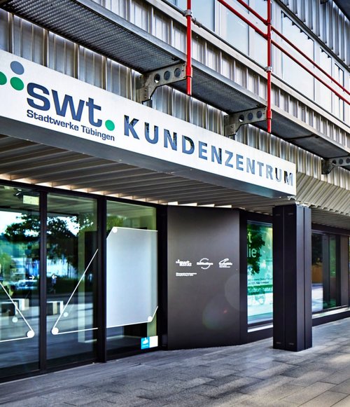 Das Bild zeigt das Kundenservicezentrum der Stadtwerke Tübingen, markiert mit dem Schriftzug "swt KUNDENZENTRUM". Die Fassade ist modern gestaltet, mit glasierten Eingängen und einem klaren, minimalistischen Design. Es gibt große Fenster, die auf eine gepflasterte Fläche vor dem Gebäude blicken. Im Hintergrund sind Bäume und andere Gebäude sichtbar, und ein Fahrradständer steht neben dem Eingang. Der Himmel ist klar und sonnig.