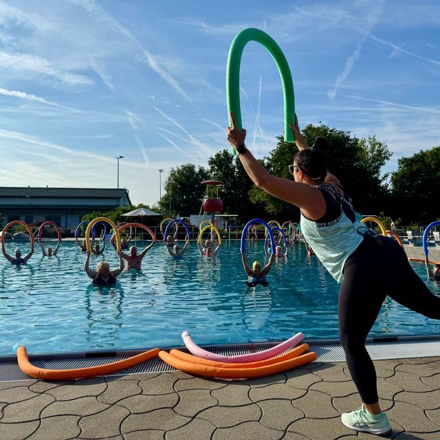Eine Übungsleiterin zeigt Bewegungen im Freibad, während Teilnehmer sich mit Schwimmreifen im Wasser bewegen.
