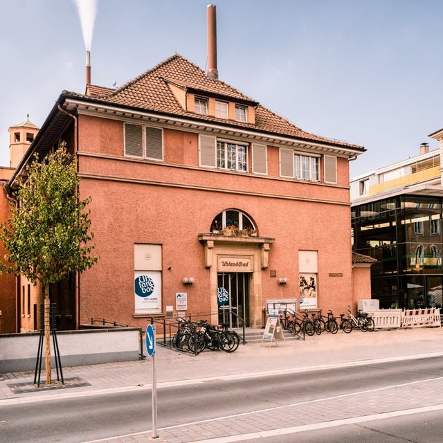 Eine Straßenansicht mit zwei Gebäuden. Auf der linken Seite befindet sich ein rotes, historisches Gebäude mit dem Namen "UhlanBad" über dem Eingang. Es gibt einen Gehweg, an dem mehrere Fahrräder abgestellt sind. Rechts daneben ist ein modernes, verglastes Gebäude zu sehen, das eine Café-ähnliche Außenterrasse hat. Im Hintergrund sind weitere mehrstöckige Gebäude sichtbar, einige Bäume sind entlang der Straße gepflanzt. Der Himmel ist klar mit einigen Wolken.
