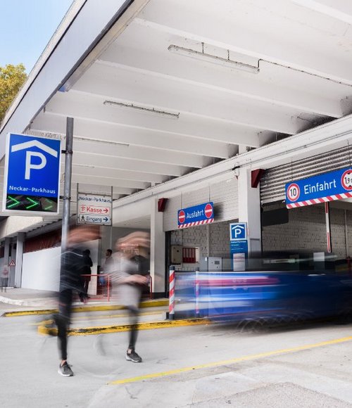 Ein Parkplatz mit einem großen "P"-Schild und der Aufschrift "Neckar-Parkhaus". Es gibt mehrere Verkehrsschilder, darunter "Einfahrt" mit einer Höhenbeschränkung von 1,9 m, "Ausfahrt", "Kasse" und "Eingang". Menschen gehen in der Nähe, während ein Auto schnell durch die Einfahrtszone fährt. Im Hintergrund sind grüne Bäume und eine moderne Architektur zu sehen.