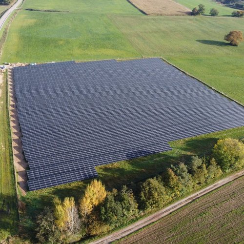 Eine große Solarenergie-Anlage, die aus vielen Solarplatten besteht, ist auf einem offenen Feld zu sehen. Das Feld ist umgeben von landwirtschaftlichen Flächen und einer Straße, die entlang der linken Seite verläuft. Im Hintergrund sind Bäume und verschiedene Grüntöne zu erkennen, die einem herbstlichen Landschaftsbild entsprechen.