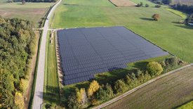 Eine große Solarenergie-Anlage, die aus vielen Solarplatten besteht, ist auf einem offenen Feld zu sehen. Das Feld ist umgeben von landwirtschaftlichen Flächen und einer Straße, die entlang der linken Seite verläuft. Im Hintergrund sind Bäume und verschiedene Grüntöne zu erkennen, die einem herbstlichen Landschaftsbild entsprechen.