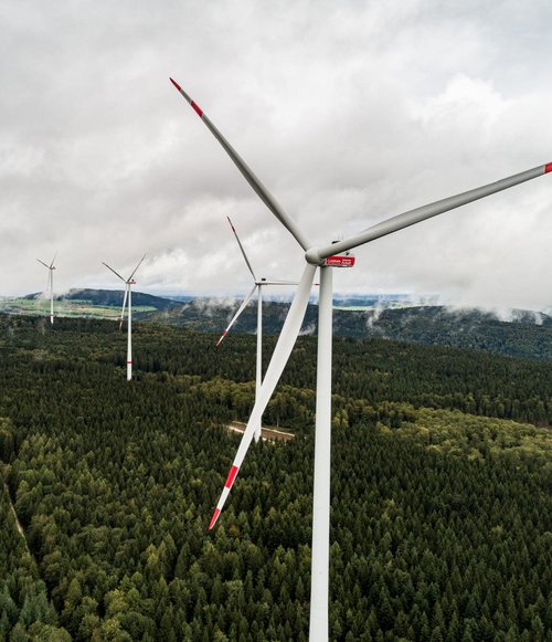 Eine Luftaufnahme von mehreren Windkraftanlagen, die zwischen dichten, grünen Wäldern stehen. Die Windräder sind von oben zu sehen, und ihre Rotorblätter sind in Bewegung. Der Himmel ist bewölkt, mit einigen Lichtreflexionen auf den Klingen. Im Hintergrund erheben sich sanfte Hügel, die von bewaldeten Flächen und Wiesen durchzogen sind.