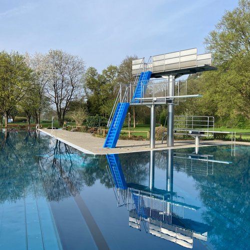 Ein Schwimmbecken mit einem Sprungturm in der Nähe. Der Sprungturm hat eine blaue Treppe und eine Plattform. Der Pool hat eine glatte, reflektierende Wasseroberfläche, umgeben von Bäumen mit frischem, grünem Laub. Im Hintergrund sind einige Bäume und eine Wiesenlandschaft zu sehen.