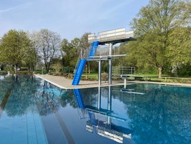 Ein Schwimmbecken mit einem Sprungturm in der Nähe. Der Sprungturm hat eine blaue Treppe und eine Plattform. Der Pool hat eine glatte, reflektierende Wasseroberfläche, umgeben von Bäumen mit frischem, grünem Laub. Im Hintergrund sind einige Bäume und eine Wiesenlandschaft zu sehen.