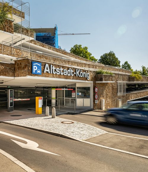Ein blaues Auto fährt in das moderne Parkhaus „Altstadt-König“ in Tübingen ein. Die Fassade besteht aus Naturstein, das Gebäude ist begrünt, und eine Schrankenanlage regelt die Zufahrt.