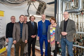 Eine Gruppe von sieben Personen steht in einem Raum mit sichtbaren Rohren und technischen Anlagen im Hintergrund. Die Personen lächeln und sind unterschiedlich gekleidet; einige tragen Anzüge, andere sportliche Kleidung. An der Wand neben ihnen hängt ein Plan, auf dem verschiedene Termine und Projekte aufgeführt sind.