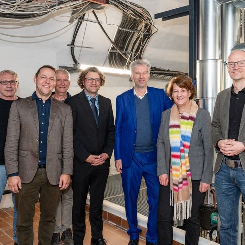 Eine Gruppe von sieben Personen steht in einem Raum mit sichtbaren Rohren und technischen Anlagen im Hintergrund. Die Personen lächeln und sind unterschiedlich gekleidet; einige tragen Anzüge, andere sportliche Kleidung. An der Wand neben ihnen hängt ein Plan, auf dem verschiedene Termine und Projekte aufgeführt sind.