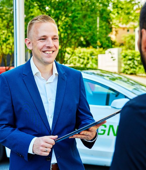 Ein lächelnder Mann in einem blauen Blazer steht an einer Ladestation für Elektrofahrzeuge und hält ein Tablet in der Hand. Im Hintergrund ist ein Elektroauto zu sehen, das teilweise im Bild ist. Ein anderer Mann, dessen Gesicht nicht vollständig sichtbar ist, steht in der Nähe und scheint mit dem lächelnden Mann zu sprechen. Die Umgebung ist grün mit Bäumen und moderner Architektur.