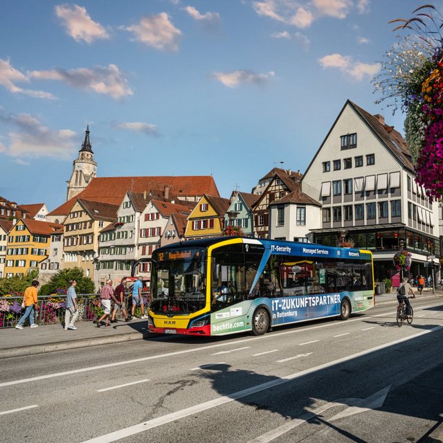 Eine Stadtstraße mit einem gelben und grünen Bus, der an einer Haltestelle hält. Im Hintergrund sind bunt bemalte Fachwerkhäuser und ein Kirchturm zu sehen. Blumenampeln hängen an den Laternen, und einige Personen gehen auf dem Gehweg. Fahrräder sind ebenfalls sichtbar, und die Atmosphäre ist hell und freundlich mit einem blauen Himmel und ein paar Wolken.