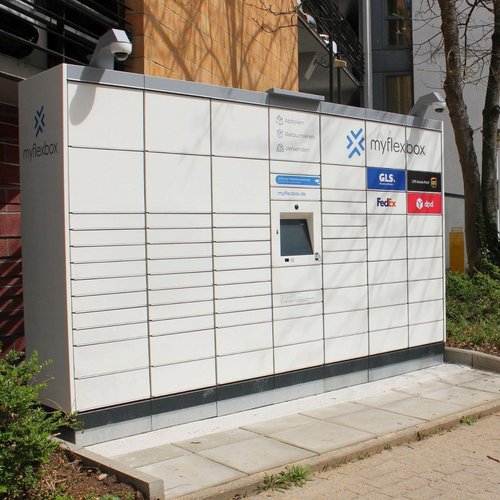 Ein Paketautomat steht an einer Straße, umgeben von Pflanzen und einem Bürgersteig. Er ist hell und modern gestaltet, mit verschiedenen Logos von Paketdiensten wie GLS, FedEx und DPD. Auf dem Automaten befindet sich ein Bildschirm und Informationen zu den angebotenen Dienstleistungen wie Abholen, Retouren und Versenden. Im Hintergrund sind Gebäude zu sehen, darunter eine Werbetafel. Ein Parkplatzschild ist ebenfalls sichtbar.