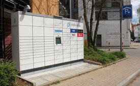 Ein Paketautomat steht an einer Straße, umgeben von Pflanzen und einem Bürgersteig. Er ist hell und modern gestaltet, mit verschiedenen Logos von Paketdiensten wie GLS, FedEx und DPD. Auf dem Automaten befindet sich ein Bildschirm und Informationen zu den angebotenen Dienstleistungen wie Abholen, Retouren und Versenden. Im Hintergrund sind Gebäude zu sehen, darunter eine Werbetafel. Ein Parkplatzschild ist ebenfalls sichtbar.