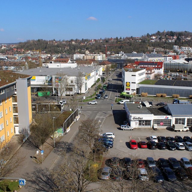 Eine Aussichtsaufnahme einer Stadtlandschaft, die moderne Gebäude und Straßen zeigt. Im Vordergrund sind mehrstöckige Wohngebäude mit gelber Fassade zu sehen, während sich im Hintergrund eine lebhafte städtische Umgebung mit verschiedenen Geschäften und Parkplätzen erstreckt. Die Straße führt zu einem Verkehrsknotenpunkt mit mehreren Fahrzeugen. Im Hintergrund sind bewaldete Hügel und weitere städtische Strukturen sichtbar, alles unter einem klaren blauen Himmel.