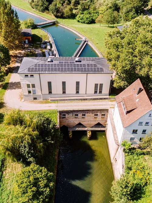 Eine Luftaufnahme zeigt eine ruhige Landschaft mit einem Wasserlauf, der von einem kleinen Gebäude mit Solarpanelen und einem weiteren, älteren Gebäude flankiert wird. Im Vordergrund sind üppiges Grün, Bäume und ein weitläufiges Feld sichtbar. Auf der linken Seite befindet sich eine kleine Sitzgelegenheit unter einem Baum. Der Wasserlauf scheint zu einem Kanal zu gehören, der sanft durch die Umgebung fließt.