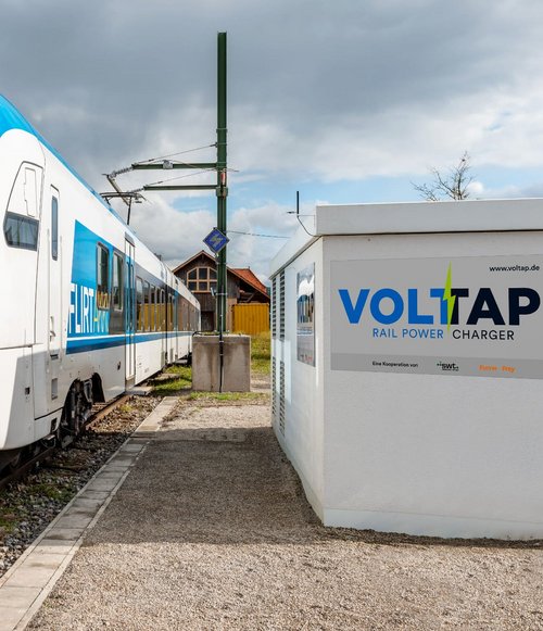 Ein weiß-blauer Zug, der von der Firma Stadler stammt, steht auf den Gleisen an einer Haltestelle. Der Zug zeigt die Anzeige "FLIRT AKKU". Neben dem Zug befindet sich ein weißes Ladegerät mit der Aufschrift "VOLTAP RAIL POWER CHARGER". Im Hintergrund sind Bäume und ein Gebäude sichtbar, und der Himmel ist teilweise bewölkt.