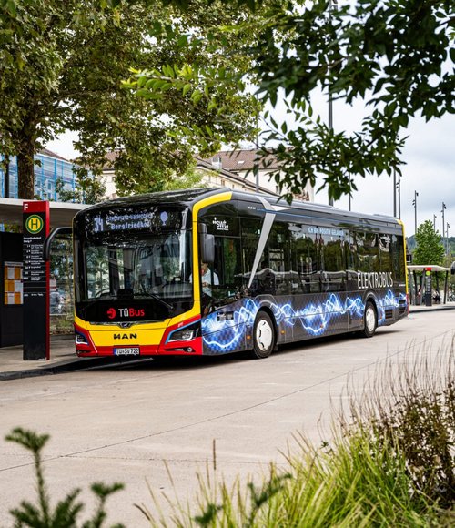 Mehrere Stadtbusse, darunter ein schwarzer Elektrobus der Linie 5 mit der Aufschrift „TüBus“, am modernen zentralen Busbahnhof Tübingen mit Haltestellen und Begrünung.