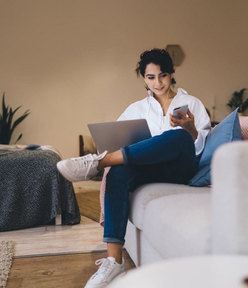 Frau sitzt auf einem Sofa mit Laptop und Smartphone, lächelnd bei der Nutzung beider Geräte.