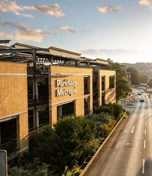 Ein Parkhaus mit dem Schriftzug "Parkhaus Metropol" ist auf der linken Seite des Bildes sichtbar. Das Gebäude hat mehrere Etagen, die teilweise mit Autos belegt sind. Im Vordergrund führt eine Straße vorbei, auf der sich Verkehrszeichen und Bäume mit grünen Blättern befinden. Im Hintergrund sind weitere Gebäude und ein leicht bewölkter Himmel zu sehen.