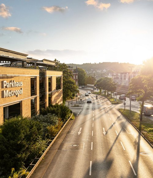 Ein Parkhaus mit dem Schriftzug "Parkhaus Metropol" ist auf der linken Seite des Bildes sichtbar. Das Gebäude hat mehrere Etagen, die teilweise mit Autos belegt sind. Im Vordergrund führt eine Straße vorbei, auf der sich Verkehrszeichen und Bäume mit grünen Blättern befinden. Im Hintergrund sind weitere Gebäude und ein leicht bewölkter Himmel zu sehen.