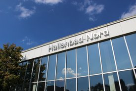 Ein modernes Gebäude mit großen Fenstern reflektiert den blauen Himmel und einige Wolken. Oben am Gebäude prangt der Schriftzug "Hallenbad Nord". Neben dem Gebäude sind einige Bäume zu sehen.