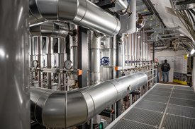 Das Bild zeigt eine industrielle Umgebung mit zahlreichen Rohren und Maschinen. Im Vordergrund sind große silberne Rohre sichtbar, die sich durch den Raum winden. Auf der linken Seite sind Messgeräte mit Ziffernblättern zu sehen, die zur Druckmessung dienen. An der Wand im Hintergrund stehen zwei Personen, die einen Plan oder ein Dokument betrachten. Der Boden besteht aus einem rutschfesten Gittermaterial, und die Decke ist mit Kabeln und Leitungen ausgestattet.