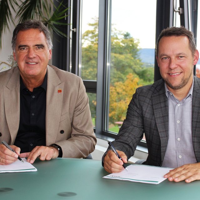Zwei Männer sitzen an einem Tisch mit einem runden, gläsernen Tisch. Sie sind in einem modernen Büro mit großen Fenstern im Hintergrund, durch die man die Natur draußen sehen kann. Beide Männer haben Schreibutensilien in der Hand und schauen in die Kamera. Der Mann links trägt einen hellen Anzug und ein schwarzes Hemd, während der Mann rechts einen karierten Blazer und ein helles Hemd trägt. Auf dem Tisch liegen Dokumente, die sie anscheinend unterschreiben möchten.