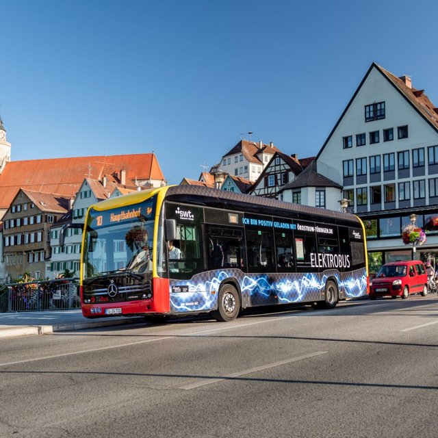 Ein Elektrobus fährt über eine Brücke in einer städtischen Umgebung. Im Hintergrund sind historische Gebäude mit roten Dächern und bunten Fenstern zu sehen. Am Rand der Straße stehen blühende Pflanzen in Töpfen. Die Sonne scheint und der Himmel ist klar und blau. Weitere Fahrzeuge und Fahrräder sind ebenfalls auf der Straße sichtbar.