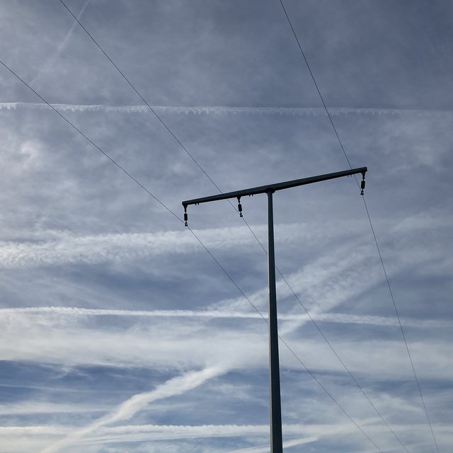 Ein Strommast steht gegen den Himmel mit mehreren Kabeln, während Wolken und Kondensstreifen in einem hellen und klaren Himmel im Hintergrund sichtbar sind. Die Wolken formen ein komplexes Muster aus Linien und Bögen.