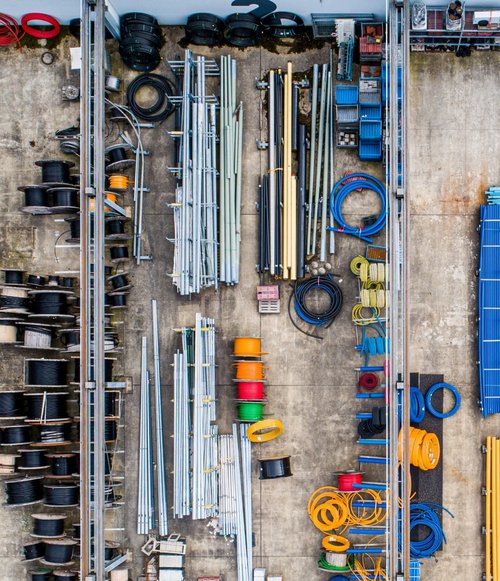 Eine Vogelperspektive auf einen Lagerraum, der verschiedene Materialien und Werkzeuge anzeigt. Auf dem Betonboden sind verschiedene Rollen und Rohre aus Metall und Kunststoff nebeneinander angeordnet. Es gibt viele schwarze, blaue, orangefarbene und gelbe Drähte oder Schläuche, die ordentlich gestapelt sind, sowie mehrere Holz- und Metallspulen. Rechts sind längliche blaue und gelbe Elemente zu sehen, die in Reihen angeordnet sind. Der Raum wirkt gut organisiert, mit klaren Linien und verschiedenen Farben, die einen visuellen Kontrast bieten.