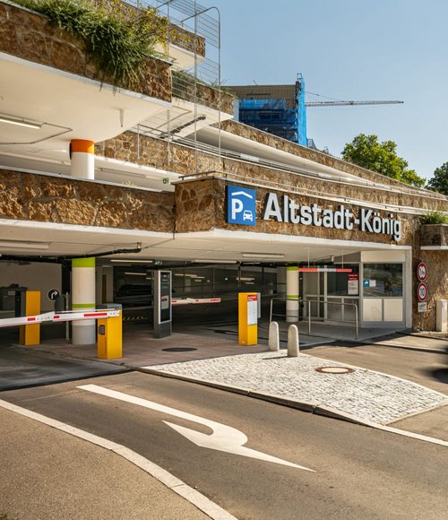 Ein Parkhaus mit dem Namen "Altstadt-König" ist im Vordergrund zu sehen. Die Fassade besteht aus Stein und ist mit Pflanzen bewachsen. Oberhalb des Eingangs sind einige sichtbare Parkebenen, die mit Geländern gesichert sind. Im Hintergrund ist ein historisches Gebäude mit einem Turm und mehreren Fenstern zu erkennen, umgeben von Bäumen und einem blauen Himmel mit einigen Wolken.