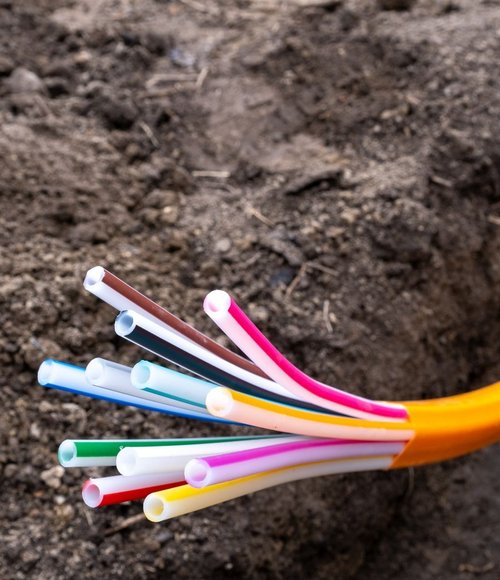 Nahaufnahme eines geöffneten orangefarbenen Schutzrohrs, aus dem mehrere bunte Leerrohre für Glasfaserkabel hervorragen, eingebettet in einem Erdgraben für den Netzausbau.