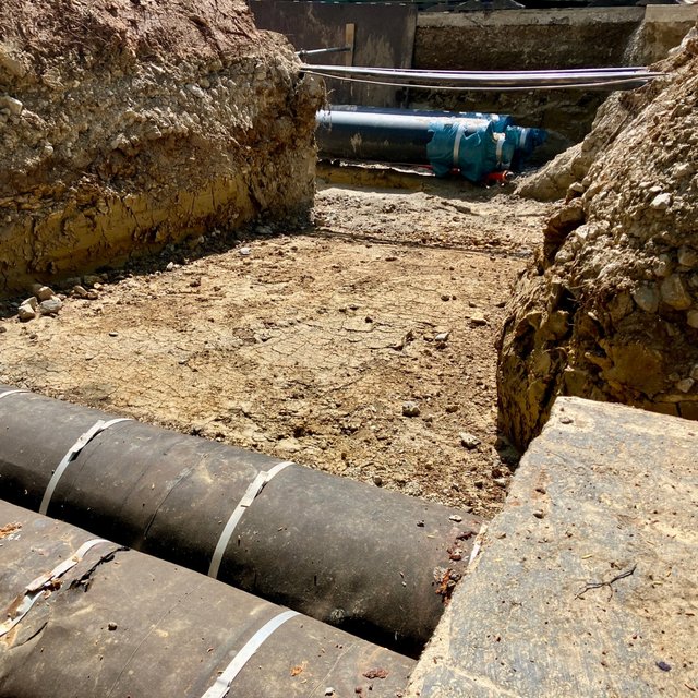 Ein Baustellenbereich mit freiliegenden Rohrleitungen. Im Vordergrund liegen zwei große, schwarze Rohre mit weißen Bändern, während im Hintergrund einige Erdhügel und eine bearbeitete Oberfläche zu sehen sind. Der Boden ist trocken und zeigt Risse, während die felsige Struktur der Erde sichtbar ist. Eine weitere Rohrleitung in blauer Folie befindet sich im Hintergrund.