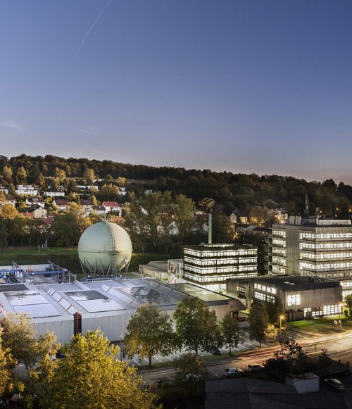 Eine weitläufige Ansicht eines modernen Industriekomplexes bei Dämmerung. Im Vordergrund sind mehrere Gebäude mit Flachdächern zu sehen, darunter ein großes, rundes, grünes Struktur mit einer Kuppel. Die Anlage ist von Bäumen umgeben und die nahen Wohngebäude sind mit roten Dächern erkennbar. Im Hintergrund erheben sich Hügel, während die Stadtlichter im Dämmerlicht beginnen zu funkeln.