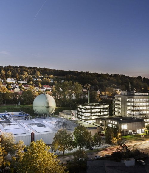 Eine weitläufige Ansicht eines modernen Industriekomplexes bei Dämmerung. Im Vordergrund sind mehrere Gebäude mit Flachdächern zu sehen, darunter ein großes, rundes, grünes Struktur mit einer Kuppel. Die Anlage ist von Bäumen umgeben und die nahen Wohngebäude sind mit roten Dächern erkennbar. Im Hintergrund erheben sich Hügel, während die Stadtlichter im Dämmerlicht beginnen zu funkeln.