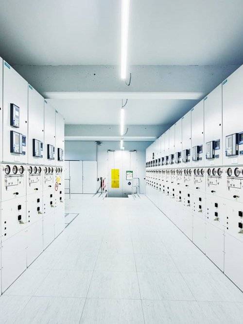 Ein modern gestalteter Kontrollraum mit mehreren weißen Schaltkästen, die an beiden Seiten des Raumes aufgestellt sind. Jeder Schaltkasten verfügt über digitale Anzeigen und Steuerungselemente. Der Boden ist hell, und die Decke ist beleuchtet von langen, weißen Lichtleisten. In der Mitte des Raumes befindet sich eine Tür, die möglicherweise zu einem weiteren Bereich führt. An einer der Wände ist ein Warnsymbol angebracht.