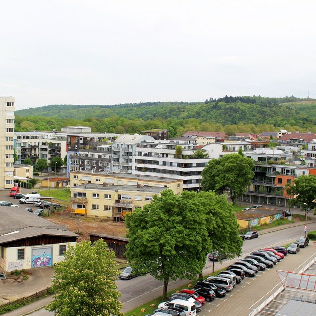In der Bildmitte steht ein mehrstöckiges, helles Wohngebäude. Daneben sind mehrere niedrigere Wohnblöcke mit unterschiedlichen Architekturstilen zu sehen. Der Hintergrund zeigt eine bewaldete Hügelkette. Im Vordergrund gibt es ein parkendes Auto mit vielen weiteren Fahrzeugen auf einem großen Parkplatz. Ein weiteres Gebäude hat eine Holzstruktur und ist teilweise mit Graffiti bemalt. Die gesamte Szene vermittelt einen städtischen Eindruck mit viel Grün um die Gebäude herum.