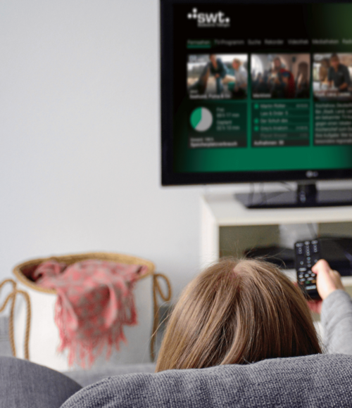 Person sitzt mit bunter Kuschelsocke entspannt auf dem Sofa und steuert mit einer Fernbedienung das IPTV-Angebot der Stadtwerke Tübingen auf einem Fernseher.