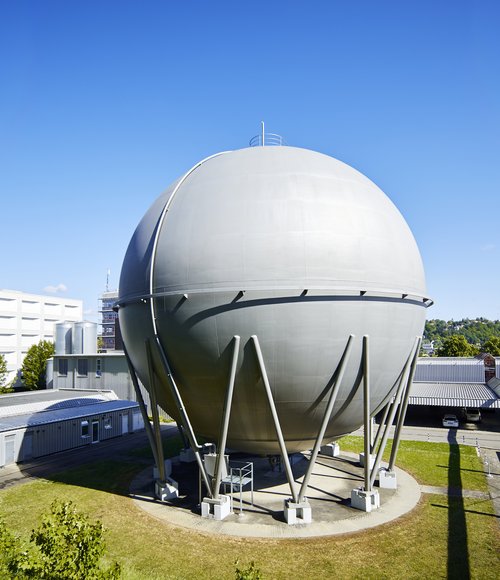 Große, silbergraue Gaskugel auf dem Betriebsgelände der Stadtwerke Tübingen bei Sonnenschein, umgeben von Zaun, Betriebsgebäuden und grüner Landschaft.