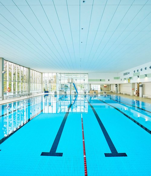 Helle Schwimmhalle im Hallenbad Nord mit großem Becken, Bahnmarkierungen, Startblöcken und großen Fensterfronten, die Tageslicht hereinlassen.