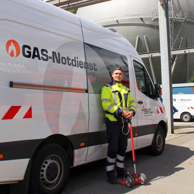 Ein Mann in einer hellen Arbeitsjacke steht neben einem Fahrzeug des GAS-Notdienstes. Er hält ein rotes Werkzeug in der Hand und lächelt. Im Hintergrund sind große silberne Tanks und ein weiteres Fahrzeug sichtbar. Die Umgebung scheint ein Industriegebiet oder ein Versorgungsbereich zu sein, mit einem klaren blauen Himmel.