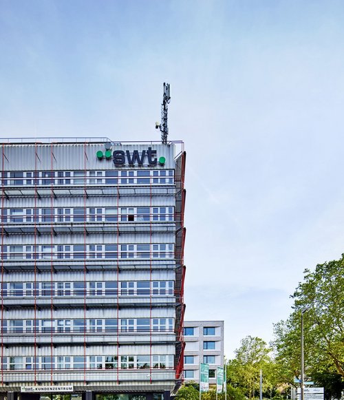 Ein modernes Gebäude mit einer transparenten Fassade, das die Aufschrift "swt" trägt. In der unteren Etage befindet sich ein Eingangsbereich mit großen Fenstern. Im Vordergrund stehen einige Sitzmöglichkeiten und ein Fahrrad. Die Straße vor dem Gebäude ist gepflastert und es gibt einige grüne Pflanzen und Bäume in der Umgebung. Der Himmel ist klar und hell.