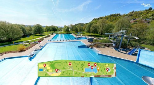 Ein weitläufiges Schwimmbad im Freien mit mehreren langen Schwimmbahnen, umgeben von grünen Bäumen und einem hügeligen Landschaftshorizont. Im Vordergrund ist ein Teil der Wasseroberfläche klar sichtbar, während im unteren Bereich eine Karte mit verschiedenen markierten Punkten zu sehen ist, die weitere Einrichtungen und Attraktionen im Bereich anzeigt. Im Hintergrund erheben sich Wasserrutschen, die zu einem Freizeitbereich gehören. Der Himmel ist blau mit wenigen Wolken.