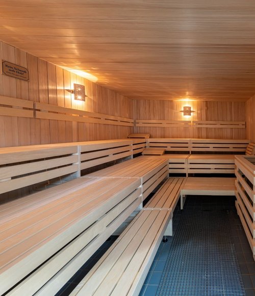 Ein einfacher, moderner Saunaraum mit Wänden und Bänken aus hellem Holz. Die Wände sind vertikal mit Holzlatten verkleidet, und es sind mehrere schlichte Holzsitze in L-Form angebracht. Die Beleuchtung besteht aus warmen Wandlampen, die sanftes Licht spenden. In der Ecke befindet sich ein Saunaofen, und der Boden ist mit blauen Fliesen und einem Teppich ausgelegt. An der Wand hängt ein Schild mit der Aufschrift „Kein Schwitzen aus Holz“.