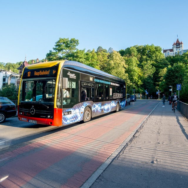 Ein Elektrobus steht an einer Haltestelle, die das Ziel "Hauptbahnhof" anzeigt. Im Hintergrund sind grüne Bäume und ein Gebäude mit einem turmähnlichen Dach sichtbar. Auf der Straße sind einige Fahrzeuge geparkt, während auf dem Bürgersteig mehrere Personen und ein Radfahrer vorbeigehen. Die Szene findet bei klarem Himmel und Tageslicht statt.