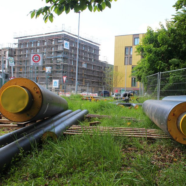 Auf dem Bild sind große schwarze Rohre mit gelben Enden zu sehen, die auf einem Gras- und Schotterboden liegen. Im Hintergrund befindet sich ein Baugerüst um ein mehrstöckiges Gebäude. Auf der gegenüberliegenden Seite sind Verkehrsschilder zu sehen, darunter ein Geschwindigkeitslimit von 30 km/h. Es gibt auch einen Sichtschutzzaun, der den Bauplatz umgibt, sowie einige Bäume und Sträucher in der Umgebung.
